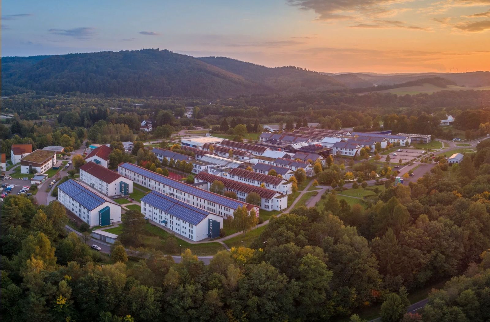 Luftbild des Umwelt-Campus Birkenfeld bei Sonnenuntergang: Gebäude mit Solardächern, ringsum Wald und Hügel.